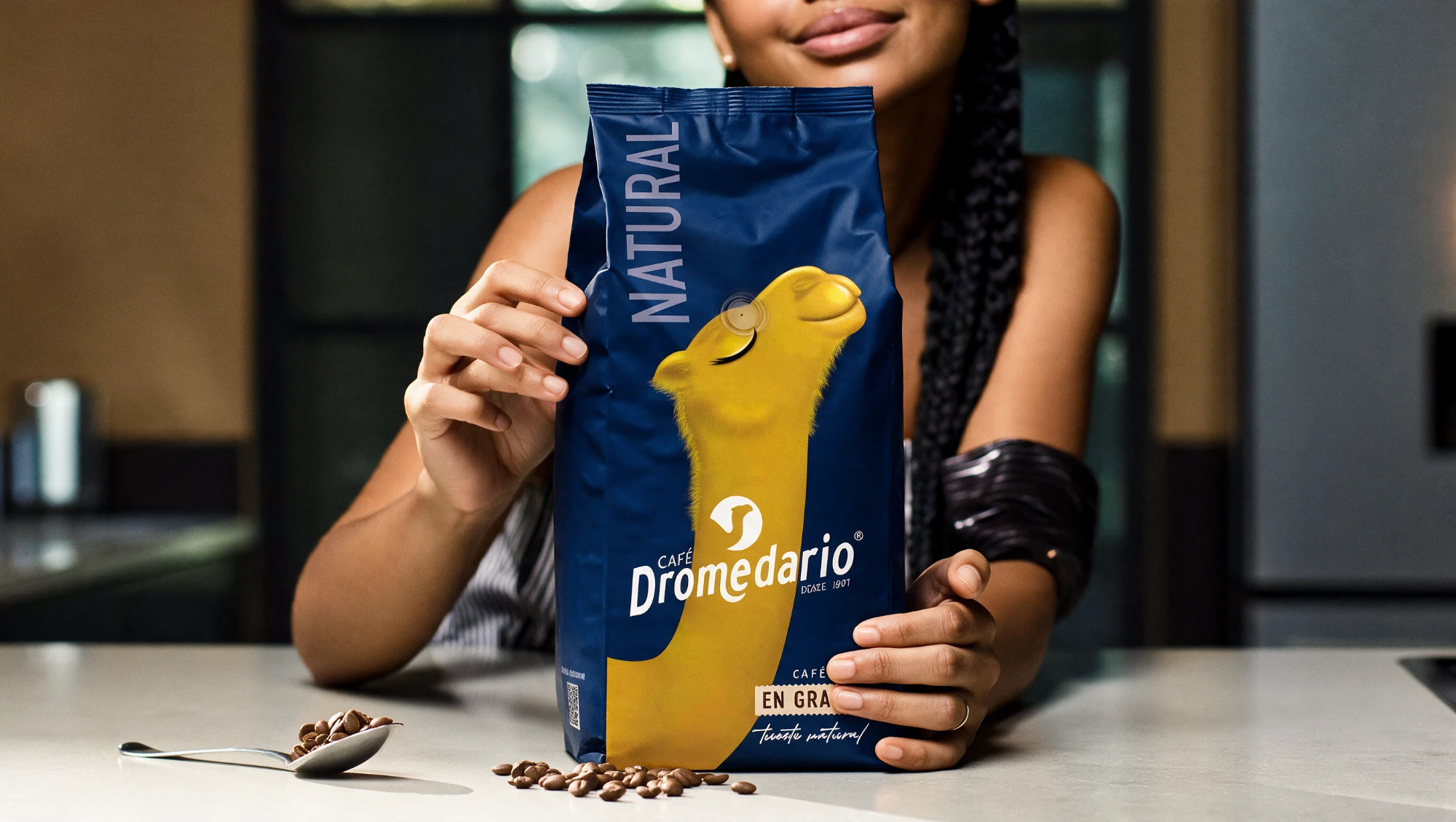 Una mujer sostiene una bolsa de Café Dromedario Natural en grano sobre una encimera, con granos de café y una cuchara